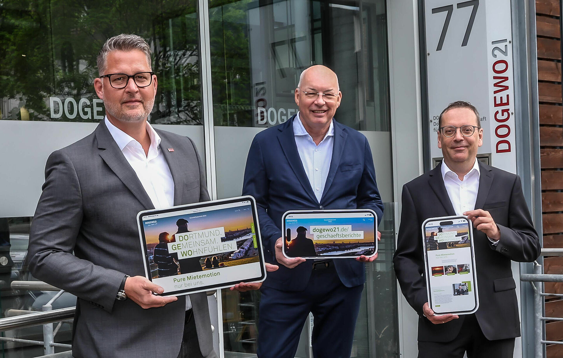 Die drei Herren der Geschäftsleitung von DOGEWO21 präsentieren auf verschiedenen digitalen Bildschirmen den Geschäftsbericht 2022